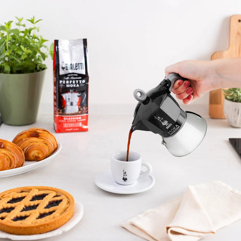 Person pouring coffee from Induction Moka Pot That Just Works On Any Hob 4 Cup Espresso Maker 5.7 Oz Black Aluminum Upper With Stainless Steel Boiler Easy Brews On Any Stove Classic Italian Coffee On Demand into a cup next to pastries.