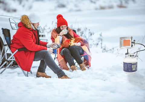 Two people seated outdoors near a GS4200EP 13,000 BTU Propane Tank Top Heater in snowy landscape with camp chairs and blanket.