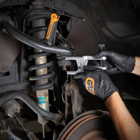 Side view of GearWrench Ball Joint Separator 3916D in use on a vehicle suspension; orange handle and silver fork.