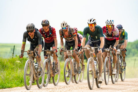 Cyclists on gravel road racing in a group, showcasing endurance riding on MTB and gravel bikes in a dirt track setting outdoors.
