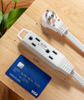 Side view of 25 Ft Space Saving Flat Plug Extension Cord With 3 Prong Ultra Thin Power Strip And 360 Degree Rotating Plug 13A 125V 1625W 16 AWG Grounded Flat Cable For Indoor Tight Spots SPT3 ETL Listed White, placed next to a credit card for size comparison on a wooden surface.