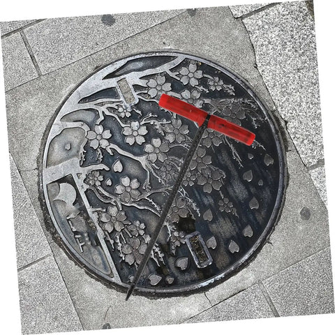Badass Heavy Duty Steel Manhole Cover Lifter Hook Built With A Nifty T Handle resting on a decorative manhole cover.