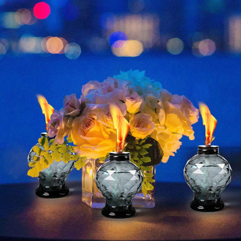 Four Pack Glass Table Torches With Citronella For Outdoor Patio And Garden on a table, surrounded by flowers, lit at night.