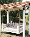 Outdoor wooden swing bed with pillows and a shingled roof structure in a grassy yard, surrounded by trees.
