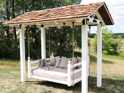 Outdoor wooden swing bed with pillows and a shingled roof structure in a grassy yard, surrounded by trees.