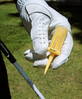 Golfer holding unbreakable yellow plastic golf tee, 3 1/8 inch, in gloved hand, against grassy background