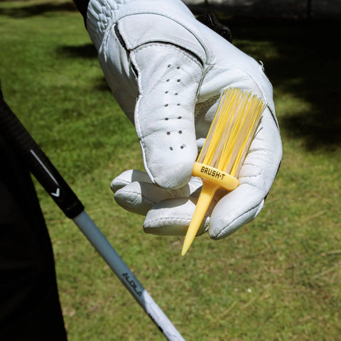 Golfer holding unbreakable yellow plastic golf tee, 3 1/8 inch, in gloved hand, against grassy background