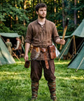 Young man in brown clothes holding a torch stands near tents, showcasing a rustic steampunk style ideal for festivals or cosplay.