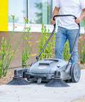 Heavy Duty Thirty Eight Inch Walk Behind Push Sweeper With Triple Side Brooms cleaning a walkway with debris around plants.