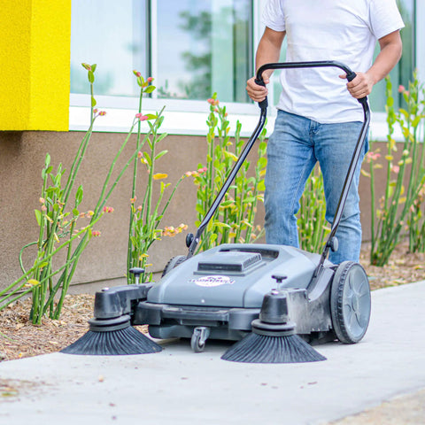 Heavy Duty Thirty Eight Inch Walk Behind Push Sweeper With Triple Side Brooms cleaning a walkway with debris around plants.