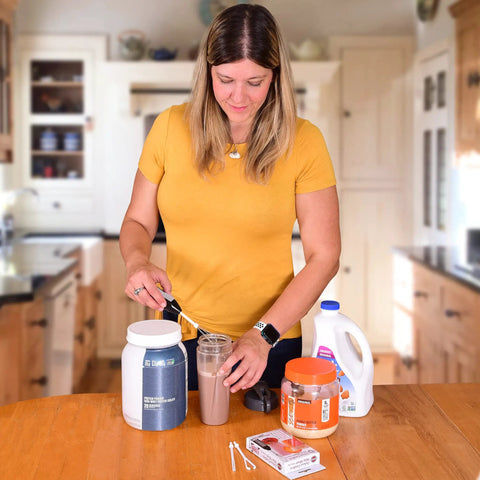 Woman in kitchen mixing protein shake with spoon, surrounded by protein powder containers and a jug of milk on the countertop.