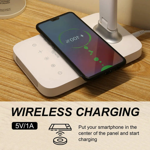 LED Desk Lamp With Wireless Charging And USB Port, smartphone charging on lamp base, touch control visible, wood desk surface