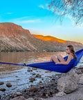 Person relaxing in Ultralight Camping Hammock 12 Oz Lightweight Blue Portable Ripstop Polyester near water, scenic mountain background.