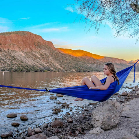 Person relaxing in Ultralight Camping Hammock 12 Oz Lightweight Blue Portable Ripstop Polyester near water, scenic mountain background.