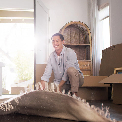 Person smiling while rolling out a rug in a sunlit room, surrounded by moving boxes and home decor, natural daylight streaming through.