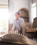 Person kneeling on the floor, smiling while rolling out a rug in a sunlit room with unpacked boxes in the background.