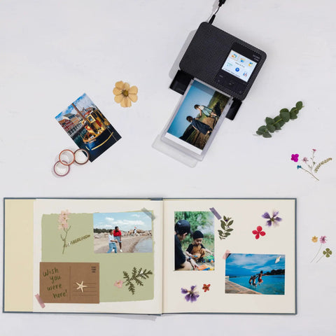 Selphy CP1500 Wireless Photo Printer In Black Ultra Portable Mini Printer printing 4x6 photos next to an open scrapbook with images