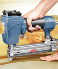 Heavy Duty Air Brad Nailer Pneumatic Nail Tool Built Tough With Durable Cast Aluminum Boasts A Slick Side Exhaust Setup For No Fuss Woodworking and Trim Work Zips 10 To 30 Millimeter Nails Home Effortlessly Without Jamming being held with one hand against wood surface.