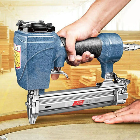 Heavy Duty Air Brad Nailer Pneumatic Nail Tool Built Tough With Durable Cast Aluminum Boasts A Slick Side Exhaust Setup For No Fuss Woodworking and Trim Work Zips 10 To 30 Millimeter Nails Home Effortlessly Without Jamming being held with one hand against wood surface.