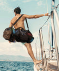 Man carrying 90L Mesh Duffle Bag For Scuba Gear And Snorkel Equipment on a boat, showcasing water sports beach essentials.