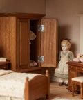 Miniature bedroom scene with dollhouse furniture, showcasing wooden wardrobe with metal hinges and a doll standing nearby.