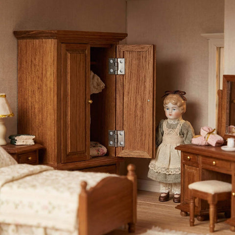 Miniature bedroom scene with dollhouse furniture, showcasing wooden wardrobe with metal hinges and a doll standing nearby.