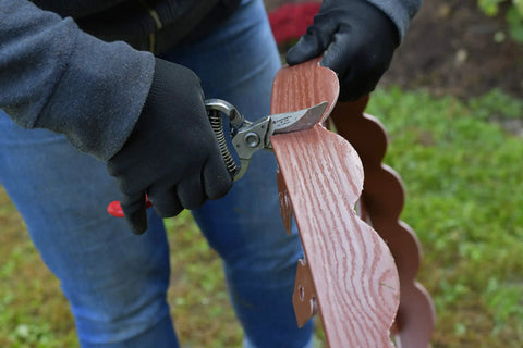 Person cutting flexible scalloped top no dig landscape edging with extra anchoring spikes in red 100 foot kit, wood grain look.