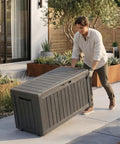 Person moving an 85 Gallon Outdoor Storage Box Resin Deck Box Waterproof UV Resistant With Reinforced Stability For Patio Garden And Poolside Striped Black Built To Last Storage For Cushions Toys And Tools on a patio.