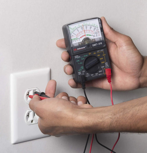 Hands using GMT 318 Analog Multimeter With Six Functions And Fourteen Ranges Reads AC And DC Volts Up To 500V on wall outlet.