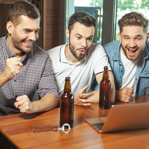 Metal bottle opener with wooden handle on wooden table; two beer bottles nearby, three men reacting in background.
