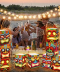 12 Pcs Hawaiian Luau Tiki Totem Gift Boxes With LED Lights For Outdoor Indoor Party Decor Vibrant Tropical Aloha Themed Gift Boxes For Tiki Bar Beach Parties And Survivor Themed Celebrations displayed at a beach party with string lights.