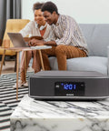 Exos Home Speaker With 30W RMS Power on marble table, living room setup in background with couple on sofa