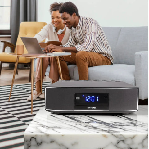 Exos Home Speaker With 30W RMS Power on marble table, living room setup in background with couple on sofa