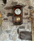Wooden wall clock on stone backdrop with pinecone weight hanging below, resembling vintage rustic home decor accent.