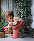 Child using a Rolling Garden Stool With Seat And Extendable Handle Tool Cart With Wheels And Extra Storage Knee Friendly Seat For Planting And Weeding Durable Steel Frame In Black next to potted plants.