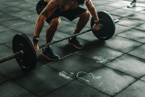 Person performing a deadlift exercise in a gym with barbell and weights on rubber flooring, wearing athletic gear.