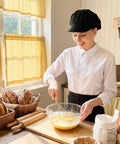 Top Shelf Three Pack Chef Hats For Women And Men With Brimmed Elastic Fit Breathable Poly Cotton Kitchen Cooking Caps For Restaurant Shifts And Weekend Cookouts worn by a chef whisking eggs in a bright kitchen.