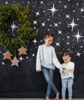 Two children standing in front of a wall adorned with the 82 Piece 3D Acrylic Mirror Star Wall Stickers Set For Ceiling And Walls, alongside a large green wreath.