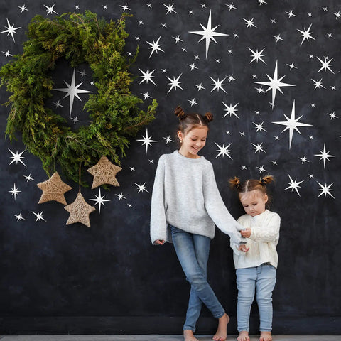 Two children standing in front of a wall adorned with the 82 Piece 3D Acrylic Mirror Star Wall Stickers Set For Ceiling And Walls, alongside a large green wreath.