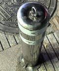 Stainless steel street bollard with lifting ring cap next to manhole cover in urban pavement setting.