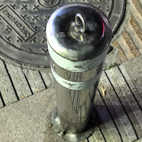 Stainless steel street bollard with lifting ring cap next to manhole cover in urban pavement setting.