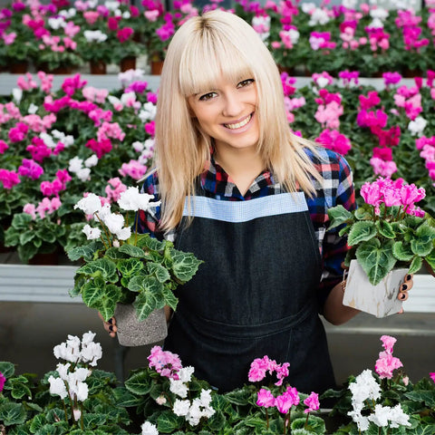 Woman in greenhouse holding pink and white potted flowers with hanging plants in background