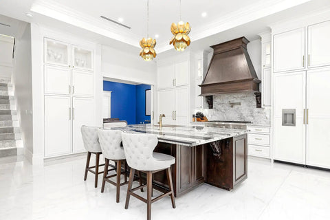 Modern white kitchen with marble island, decorative chairs, and brown vent hood. Elegant lighting and cabinets in background.