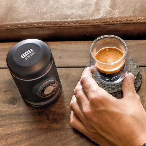 Picopresso Portable Espresso Maker Bundle With Protective Case For Pro Level Coffee On The Go Ultra Fine Grind Compatible Manual Travel Espresso System Delivers Creamy Shots And Dense Flavor next to hand holding espresso cup on table.