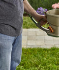 Man holding Hand Powered Lawn Spreader For Grass Seed Fertilizer Salt Ice Melt With Adjustable Arm Support in garden