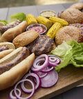 Outdoor BBQ spread on a wooden board including grilled sausages, hamburgers, lettuce, sliced onions, and corn.
