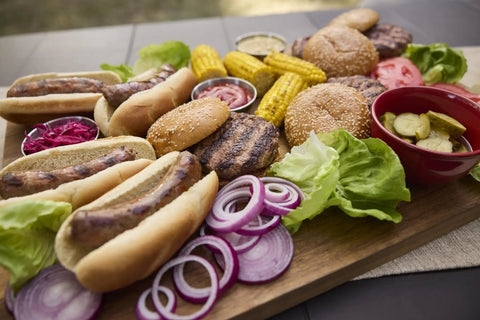 Outdoor BBQ spread on a wooden board including grilled sausages, hamburgers, lettuce, sliced onions, and corn.