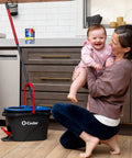 Woman holding baby in kitchen next to RinseClean Spin Mop And Bucket System For Clean Water Mopping Lets You Clean With Clean Water From Start To Finish Removes 99 Percent Of Bacteria With Just Water Built In Foot Pedal For Hands Free Wringer And Easy Rinse.