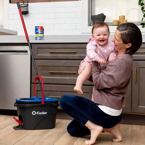 Woman holding baby in kitchen next to RinseClean Spin Mop And Bucket System For Clean Water Mopping Lets You Clean With Clean Water From Start To Finish Removes 99 Percent Of Bacteria With Just Water Built In Foot Pedal For Hands Free Wringer And Easy Rinse.