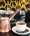 IST Series Thick Solid Copper Engraved Cezve For Stovetop Turkish Greek Arabic Coffee Pot beside full coffee cup in cafe setting